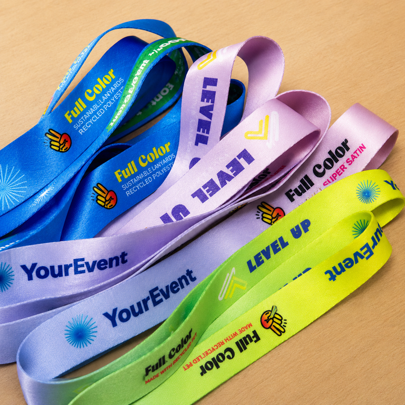 Four colorful recycled satin lanyards in blue, lavender, light purple, and neon yellow-green, fanned out on a light wood surface. Each lanyard features 'Full Color' text, a recycling symbol, and custom event text like 'YourEvent' or 'LEVEL UP'.