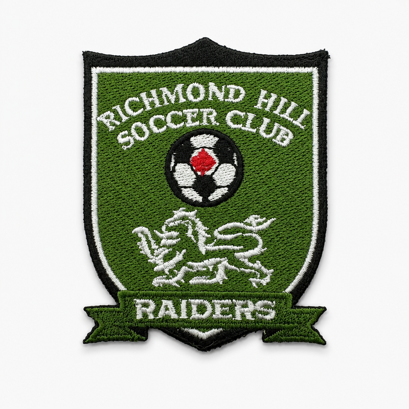 An embroidered shield-shaped patch for Richmond Hill Soccer Club, featuring a green background, white and black text, a soccer ball with a red center, a white lion rampant, and a green ribbon banner reading 'Raiders' at the bottom.