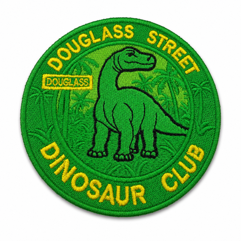 A circular embroidered patch featuring a green dinosaur silhouette in the center, surrounded by yellow text reading 'DOUGASS STREET' at the top and 'DINOSAUR CLUB' at the bottom, with a small 'DOUGASS' label on the left. The patch is green with yellow lettering and a black outline.