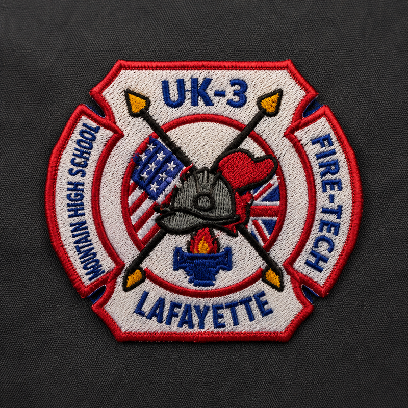 An embroidered fire department patch shaped like a Maltese cross, with the text 'UK-3', 'MORNING HIGH SCHOOL', 'LAFAYETTE', and 'FIRE-TECH' around the design. The center shows a firefighter helmet, heart, American flag, Union Jack, and a hose coil on a dark fabric background.