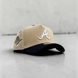 A beige baseball cap with a dark navy curved brim, featuring a white embroidered Atlanta Braves 'A' logo on the front and a team patch on the side, resting on a gray concrete surface.