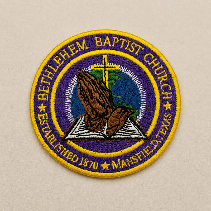 A circular embroidered patch for Bethel Baptist Church, Mansfield, Texas, featuring praying hands over an open Bible with a cross and globe motif, bordered by yellow text on a purple and yellow background.