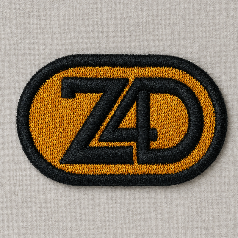 An embroidered oval patch featuring the letters 'Z4D' in black outlined embroidery over a mustard yellow background, shown on a light gray textured fabric.