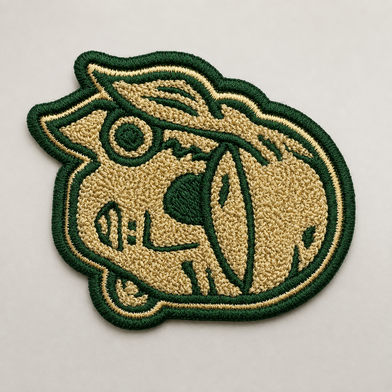 An embroidered patch featuring a stylized cartoon character head in beige and dark green, photographed on a plain light background.