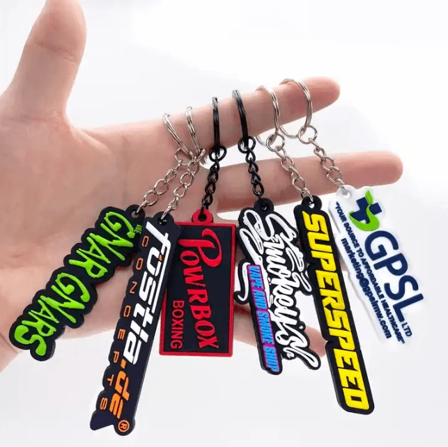 A hand holding five colorful custom rubber keychains with metal split rings and chains, featuring various racing and sports-themed designs like 'Toshiba' and 'Superspeed'.